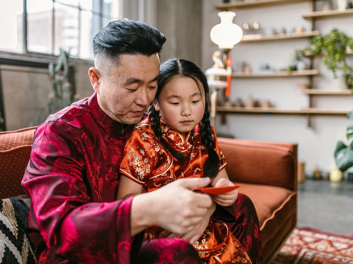 A man in a red silk tang suit and a young girl in braids and a red-orange brocade dress look at a red envelope on a sofa.