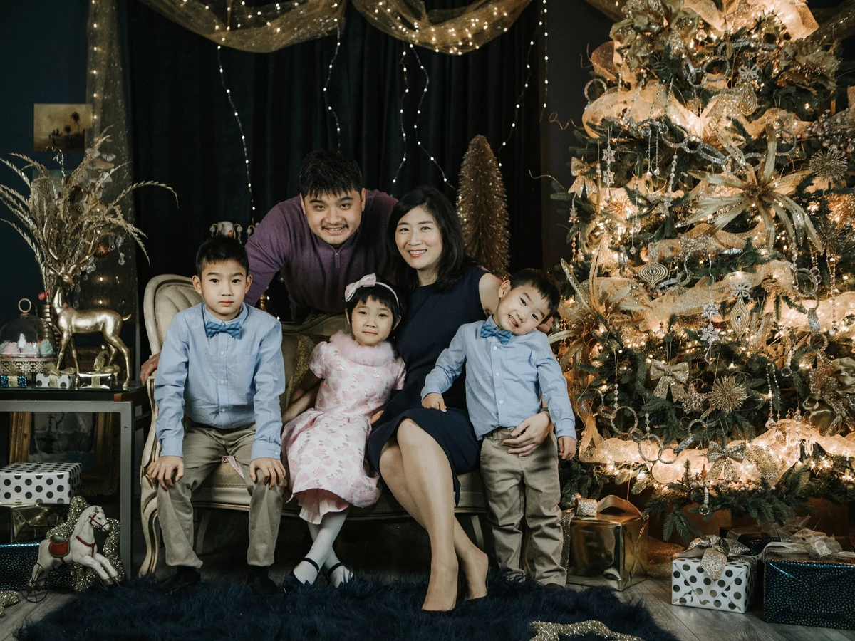 Two adults and three children pose on a sofa beside a Christmas tree with gold and white lights and wrapped presents.