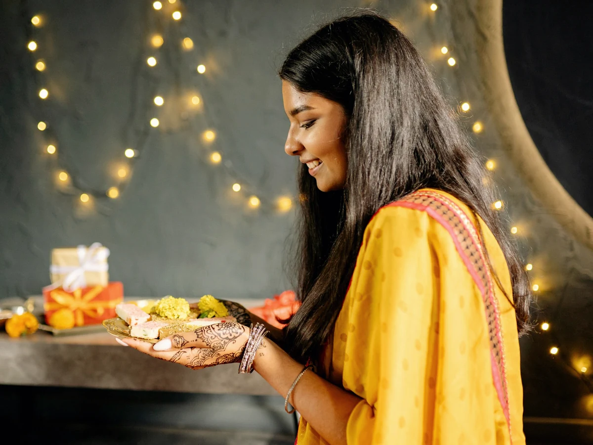 A woman in a yellow outfit with henna on her hands holds a plate of sweets in front of warm string lights.