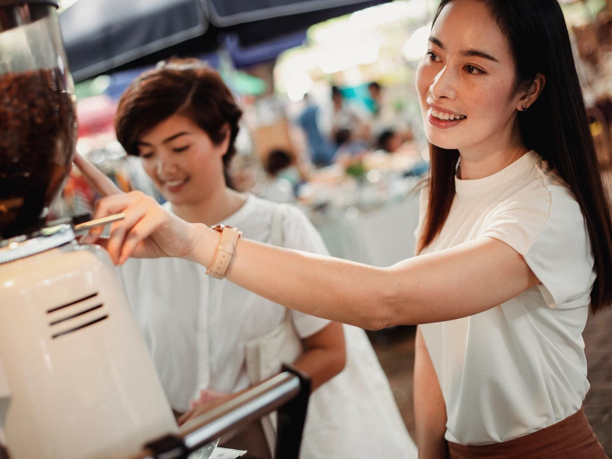 Two women smile at a street café counter as one reaches for the coffee grinder.
