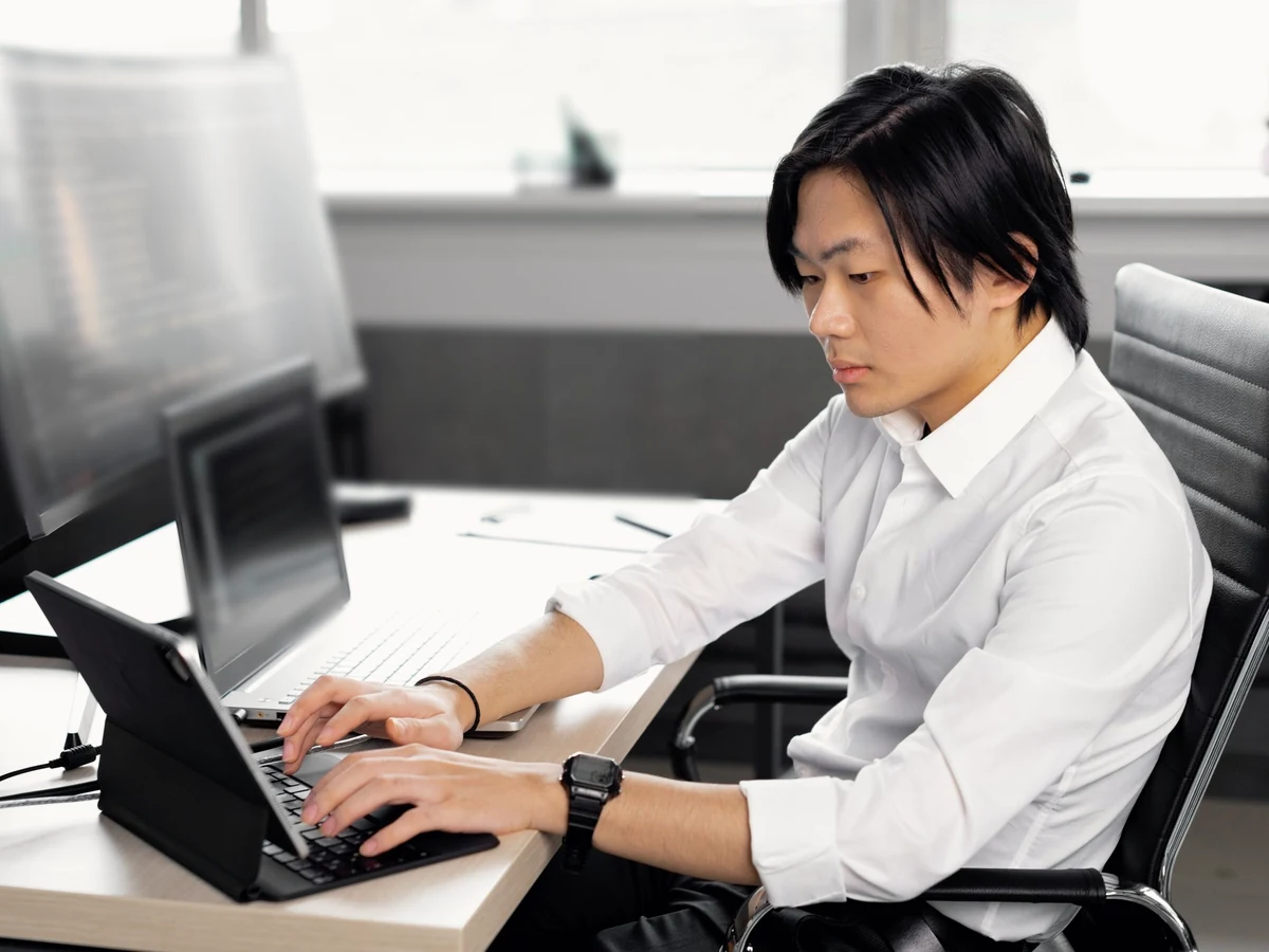 A young man in a white shirt types on a tablet keyboard at an office desk, with a laptop and a large monitor beside him.