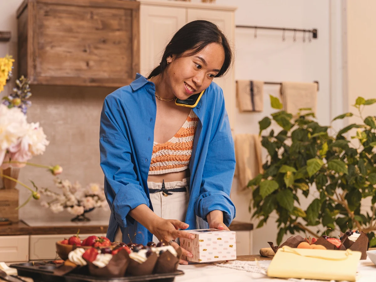 A woman in a blue shirt talks on her phone while packing pastries into a gift box at a kitchen counter.