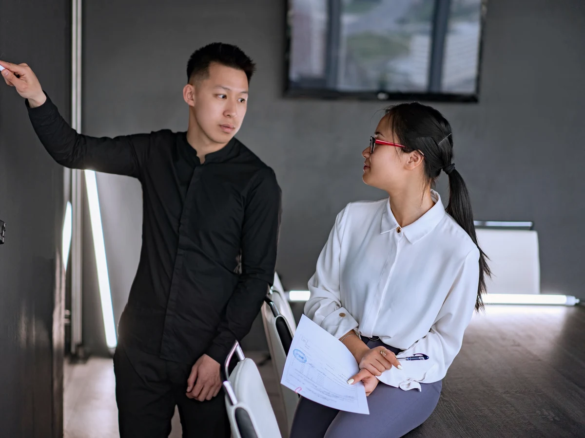 A man in a black shirt points at a dark wall board while a woman in red glasses and a white blouse holds papers and looks on.
