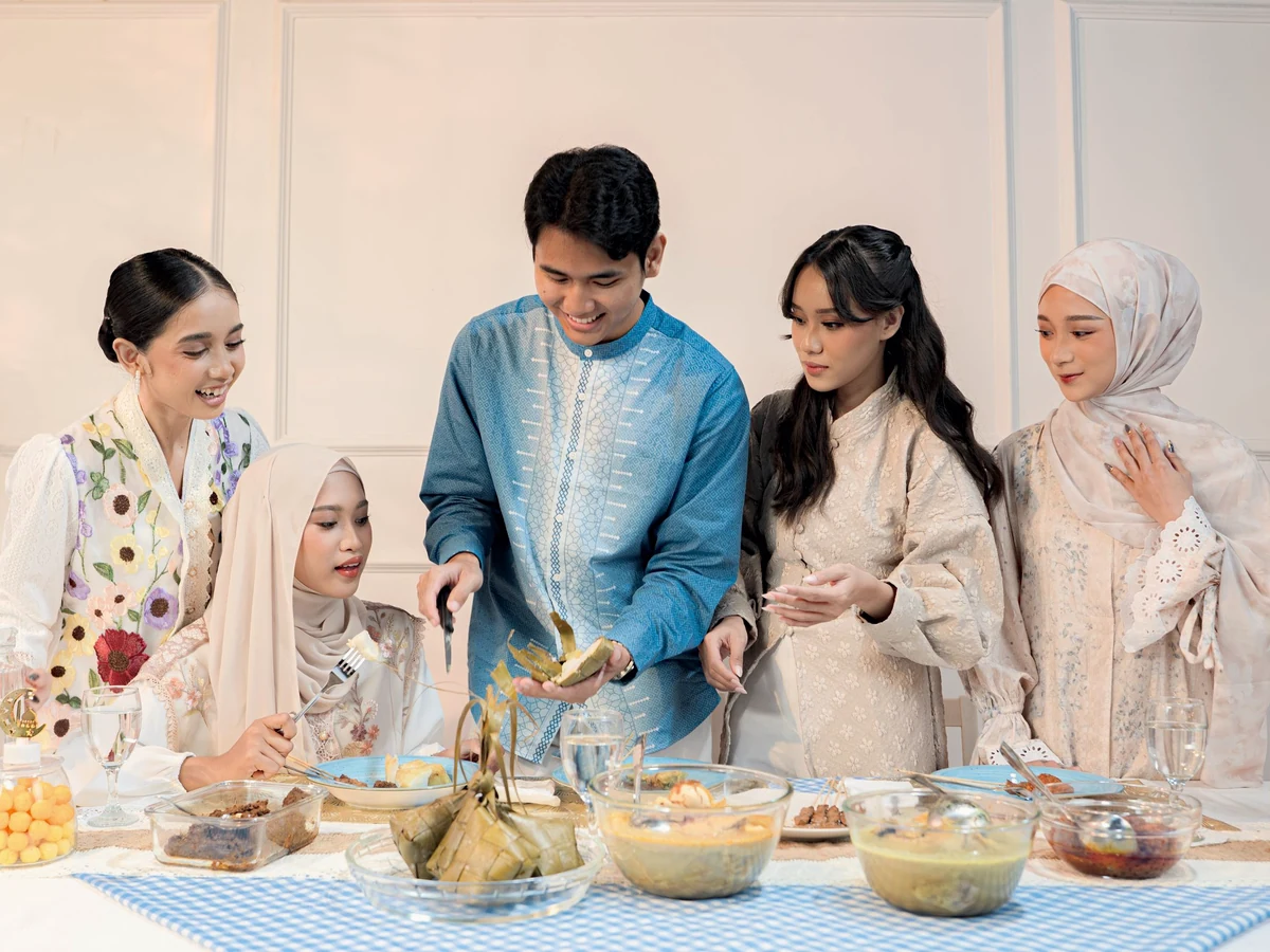 Five young adults in baju Melayu, baju kurung and hijabs smile around a table of festive dishes and ketupat.