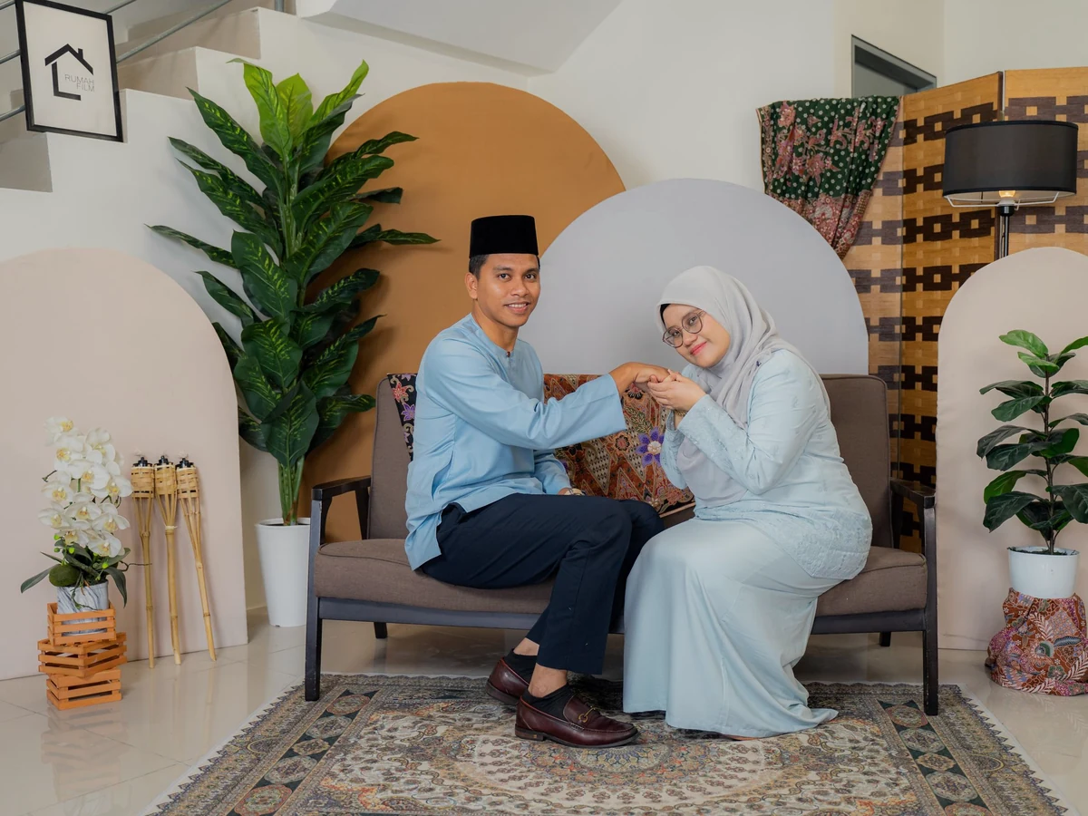 A couple in Raya clothes, he in a blue baju Melayu and songkok, she in a light-blue hijab, exchange salam on a sofa.