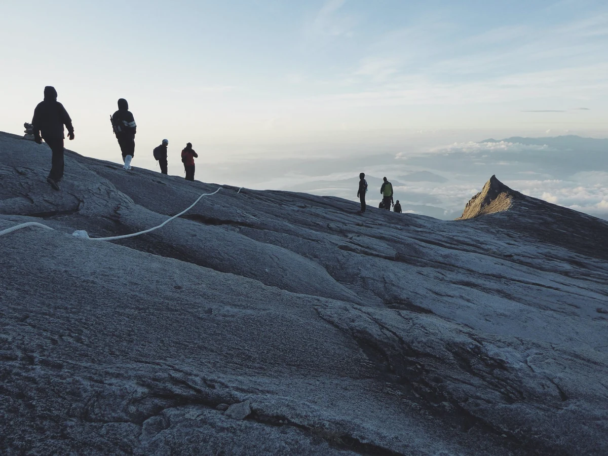 A line of hikers in silhouette crosses a bare granite mountain plateau at dawn, above a sea of clouds.