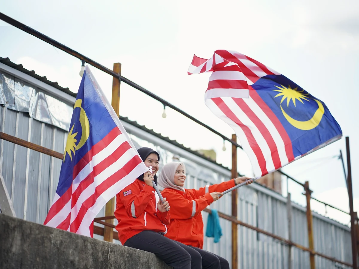 Two women in red jackets and hijabs sit on a concrete ledge waving two large Malaysian flags under an overcast sky.