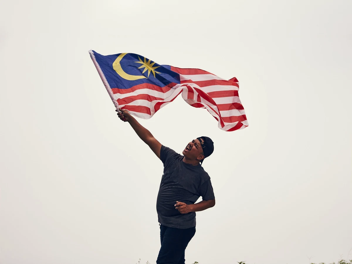 A man in a dark cap and T-shirt waves a large Malaysian flag overhead against a pale sky.
