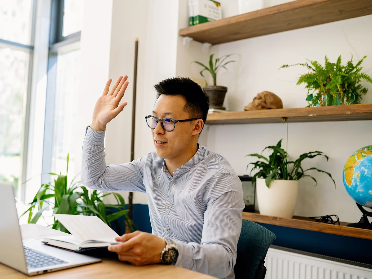 A man with glasses raises his hand at an open laptop, holding a notebook, in a home office with plants and a globe.