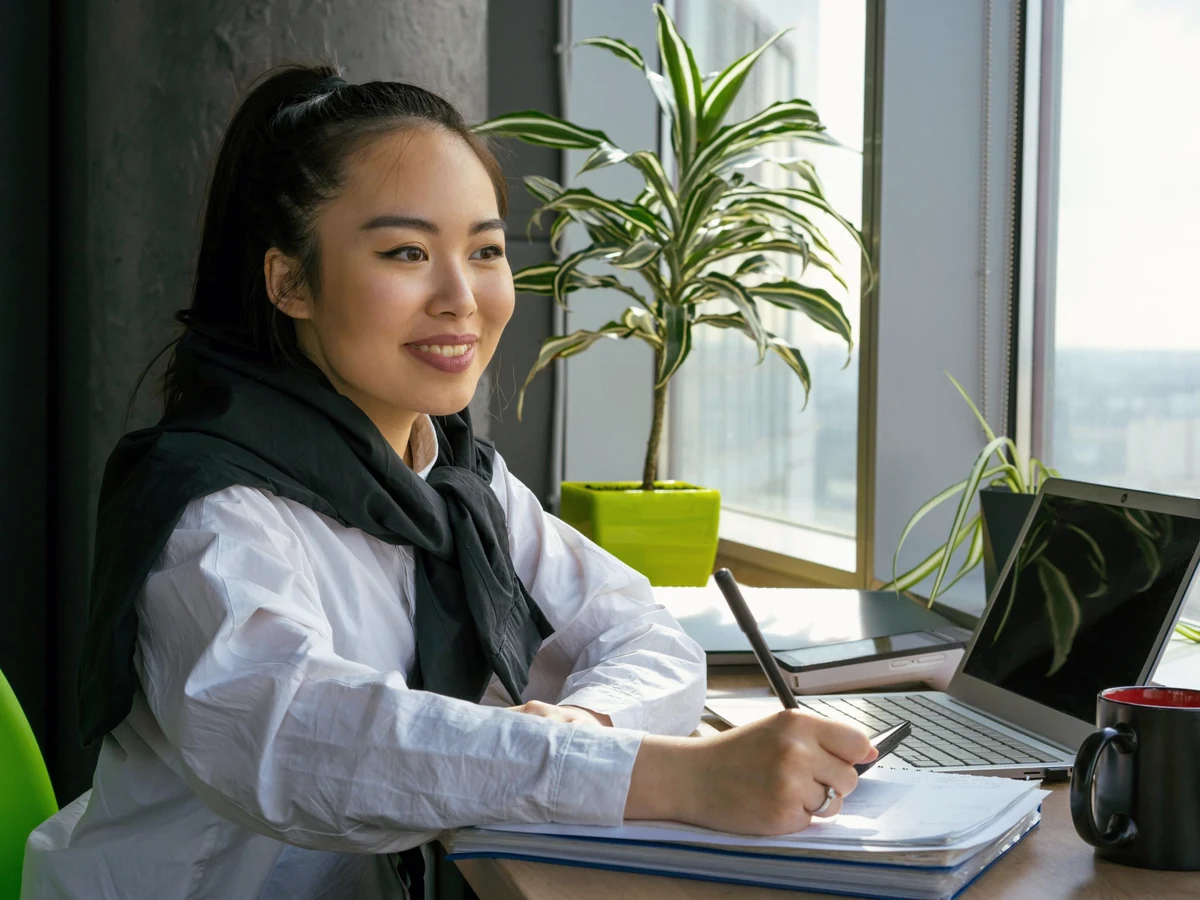 A smiling woman with a dark scarf writes notes at a desk with a laptop, beside a large window and potted plants.