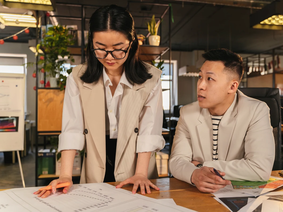 A woman in glasses leans over papers on a table as a man in a beige blazer looks on in an open-plan office.