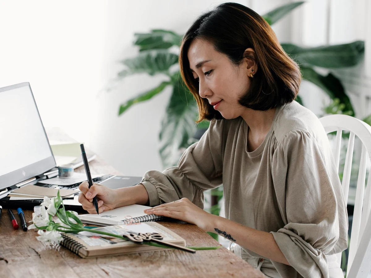 A woman with shoulder-length hair writes in a notebook at a wooden desk beside an open laptop, white flowers and a plant.