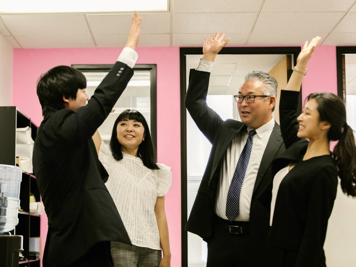 Four people in business clothes celebrate in a bright office with pink walls, three raising their hands for a high-five.